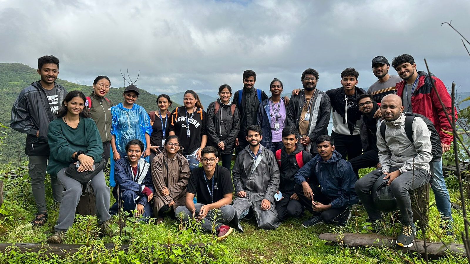 Students in raincoats on a green hilltop under monsoon clouds during a Lavasa Trekkers trek