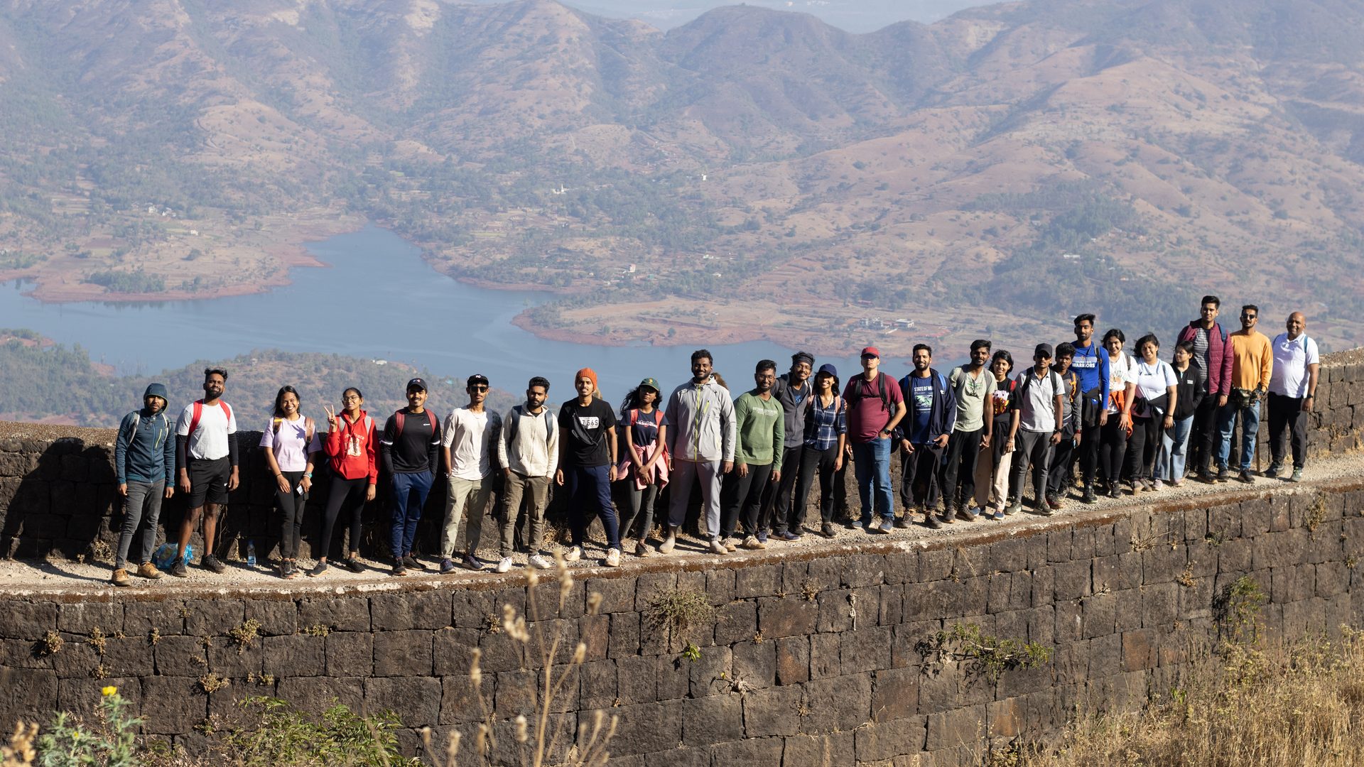 Lavasa Trekkers members lined up along the stone rampart of Torna Fort, with hills and a lake in the valley below
